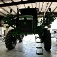 Green John Deere sprayer in a farm shed, viewed from the front with large tires and ladder visible