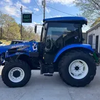 Blue New Holland tractor parked on a driveway beside a building, side view