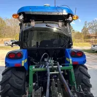 Rear view of a blue tractor with large tires and green equipment attached in a rural yard