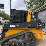 Yellow and black John Deere 325G compact track loader beside a building on a muddy lot