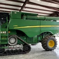 Green combine harvester with tracks and large yellow wheel parked in a barn