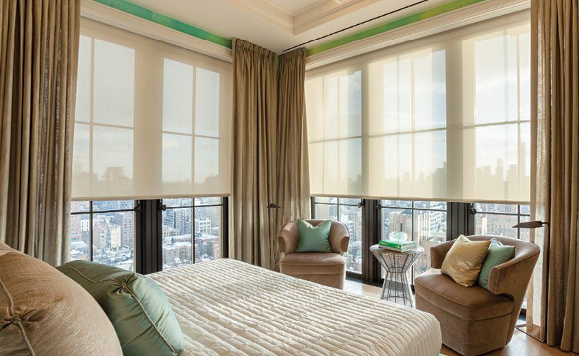 Earth toned bed, chairs, drapery and roller shades over looking a city