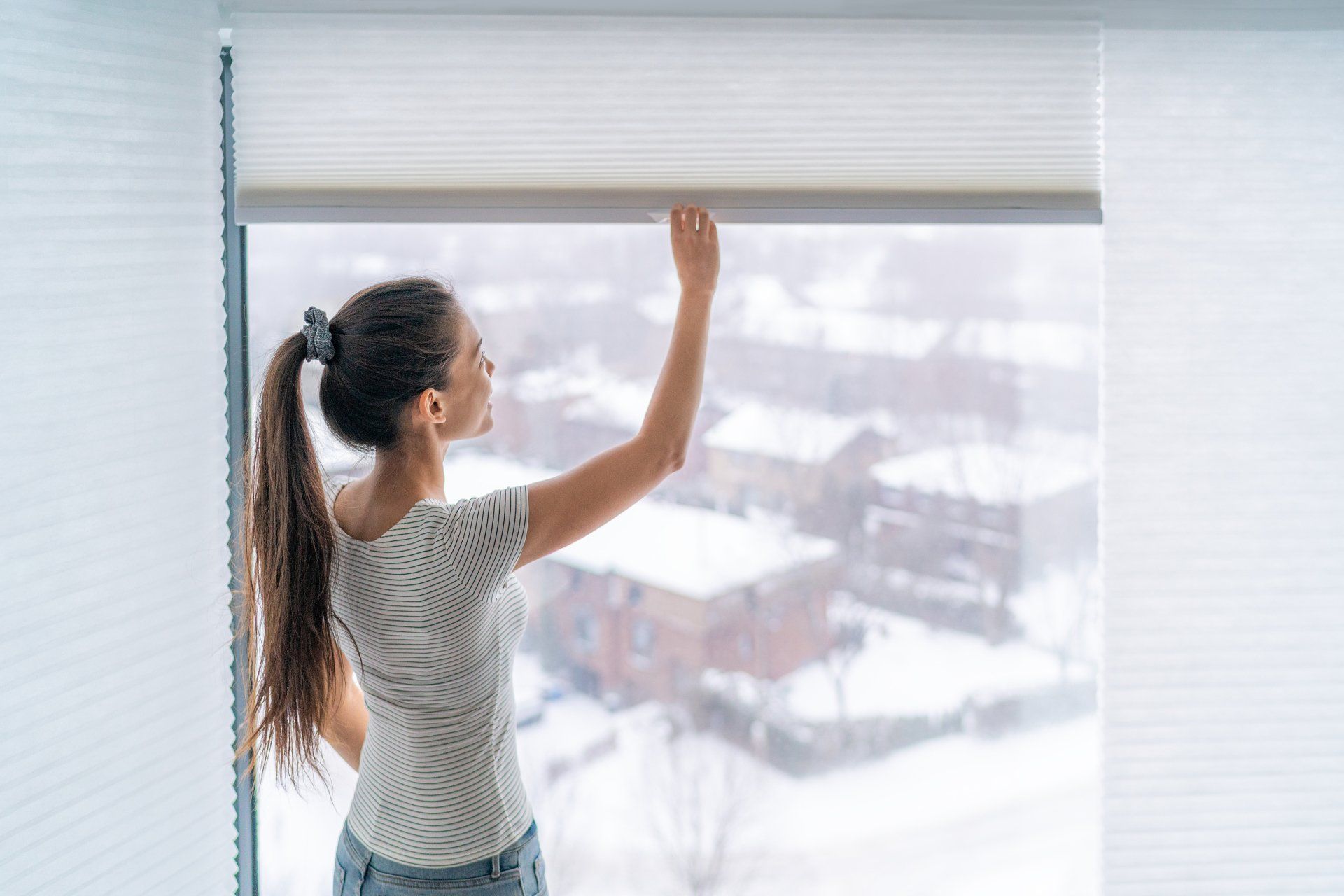 Woman closing white window shades overlooking snowy buildings