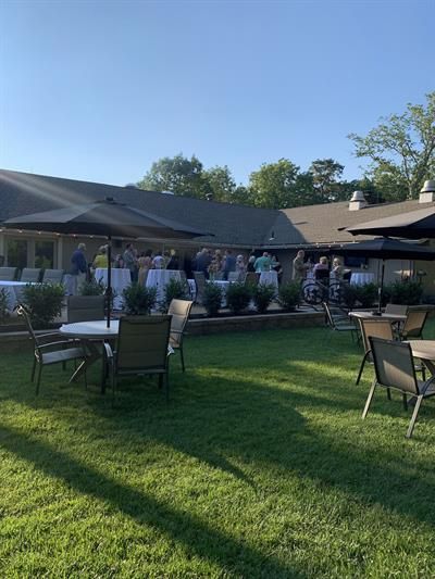 A group of people are sitting at tables and chairs in a grassy area in front of a house.