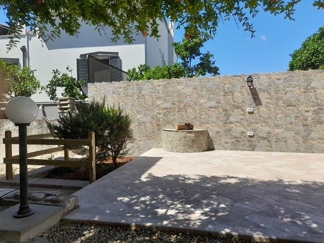 Patio esterno con muro in pietra, pavimento in cemento e pozzo rotondo. Lampione sulla sinistra, cielo azzurro.