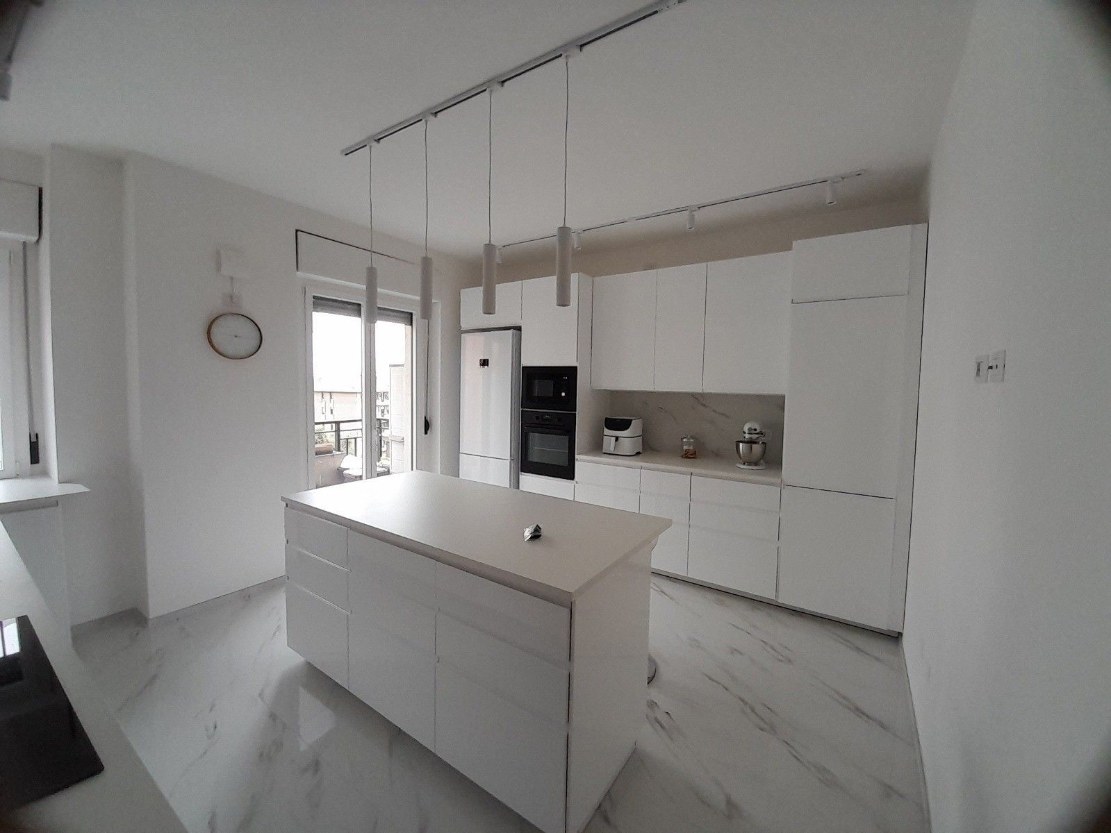 Cucina bianca con isola, mobili, elettrodomestici e lampade a sospensione.