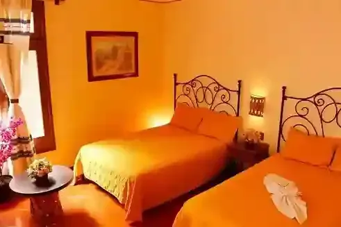 Una habitación de hotel con dos camas, ropa de cama naranja, una mesita de noche con lámpara, una mesa redonda pequeña.