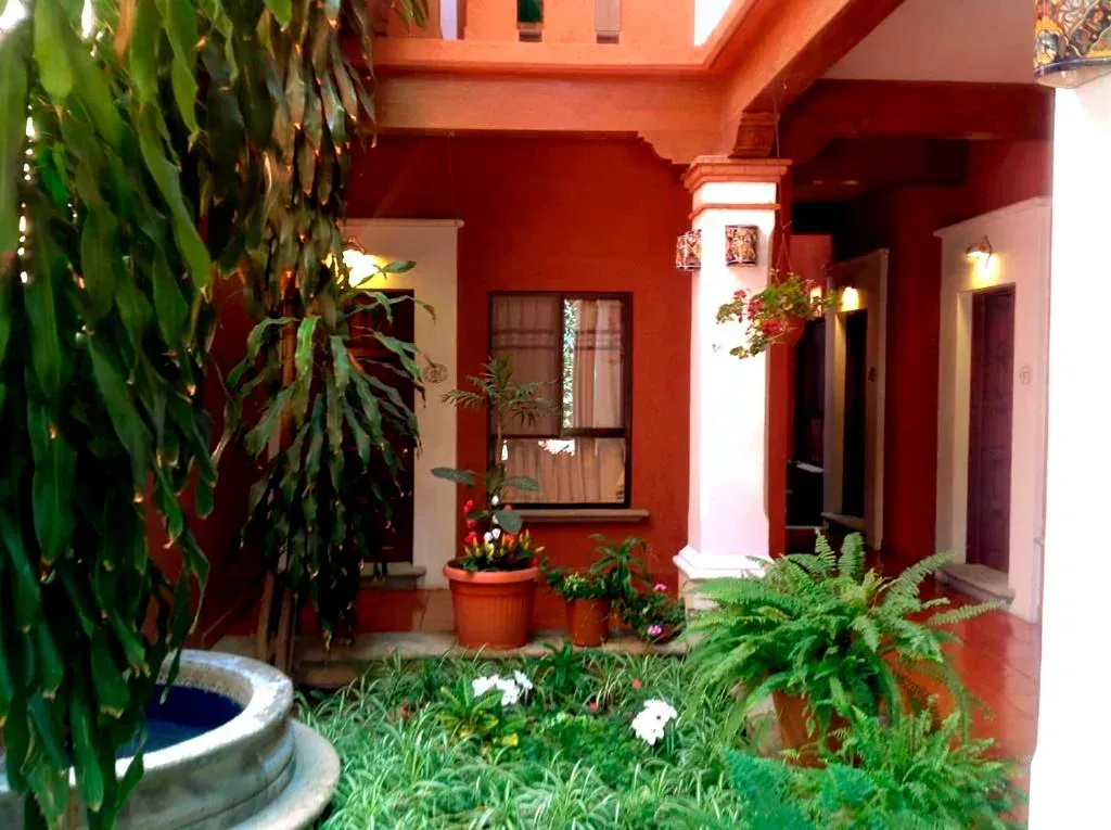 Un patio exuberante con una fuente de piedra, plantas frondosas, paredes rojas y pasillos abovedados.
