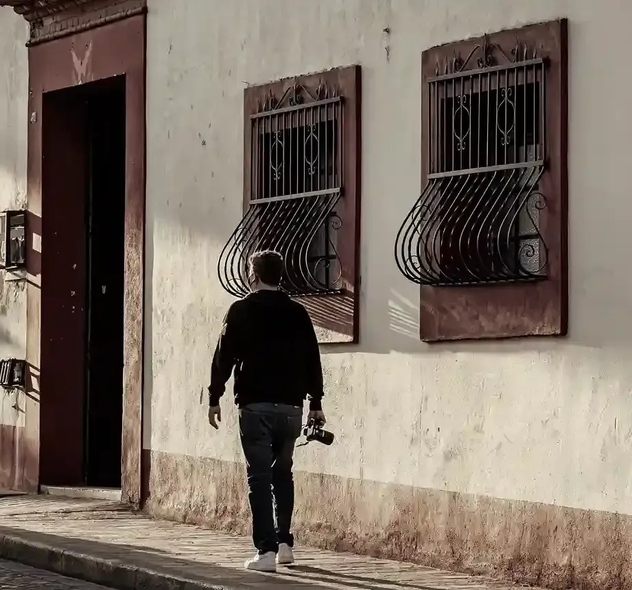 Una persona vestida con una chaqueta negra y vaqueros camina junto a una pared beige.