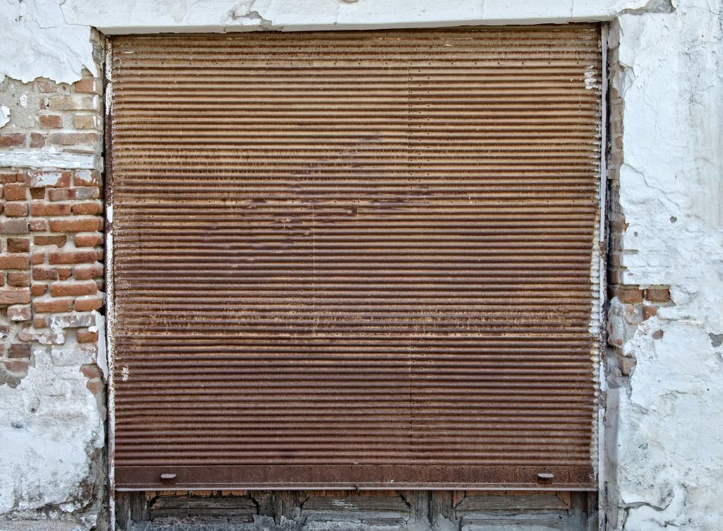 Laser clean rusty metal security shutter covers an opening in a brick wall with peeling white paint.