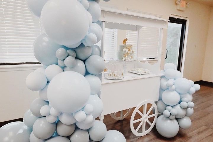 A white cart is decorated with blue balloons for a baby shower.
