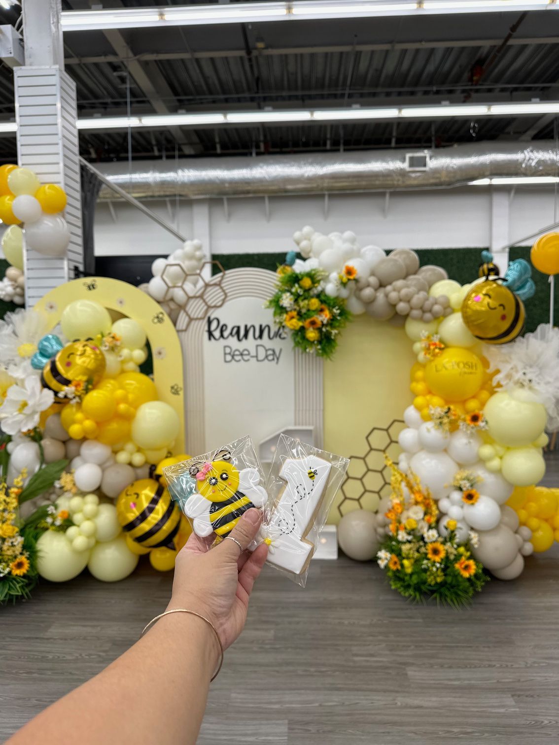 A person is holding a cookie in front of a bee themed balloon arch.