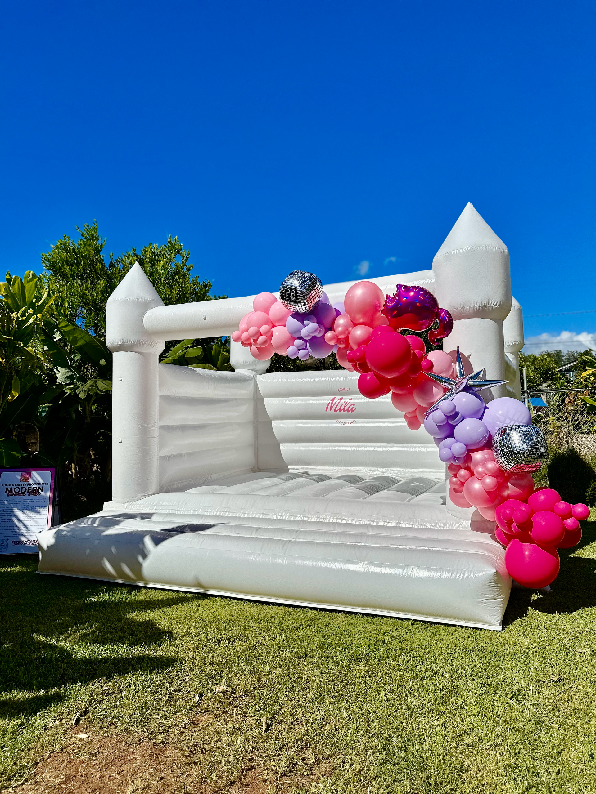 A white bouncy castle is decorated with pink and purple balloons.
