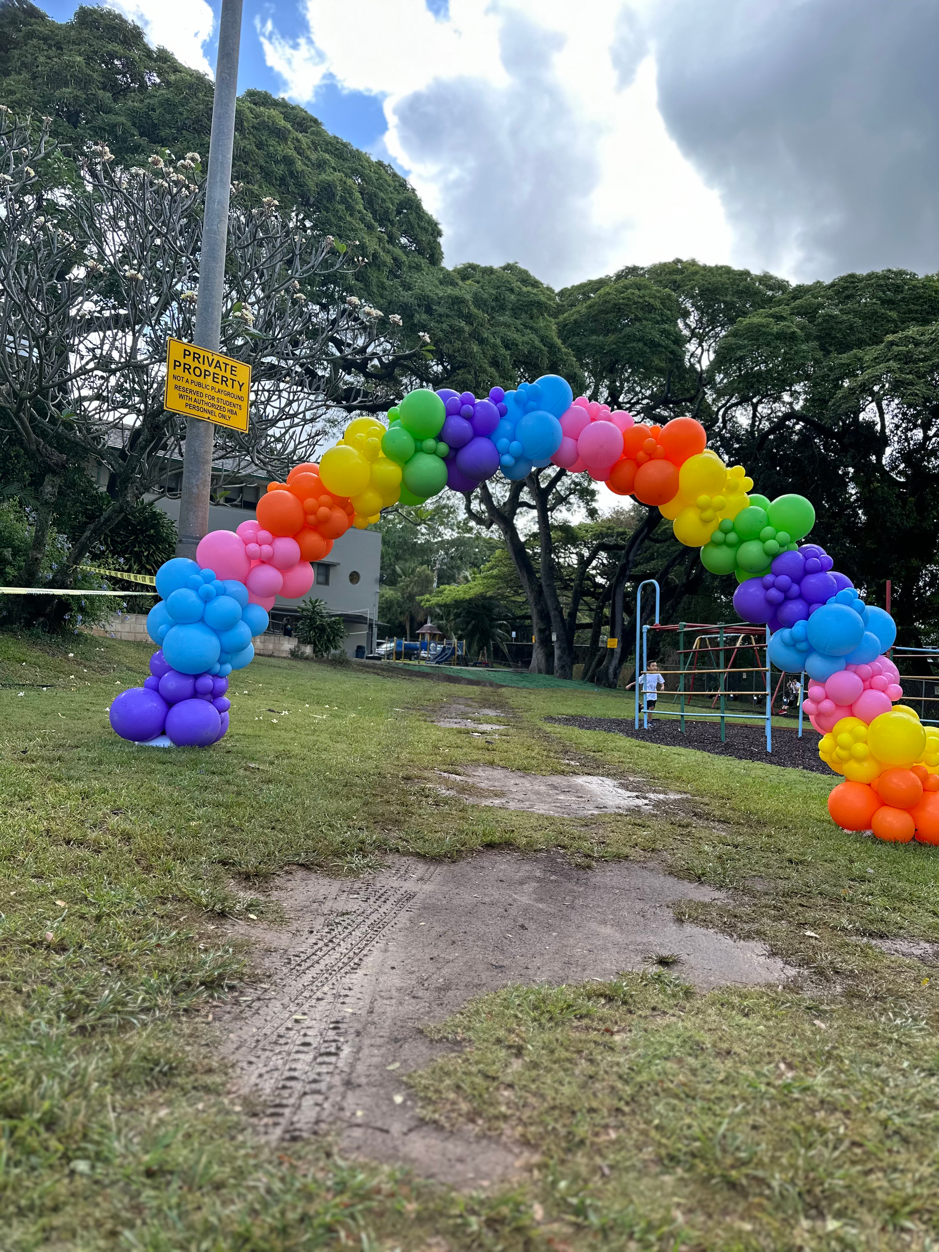 A rainbow colored balloon arch is in the middle of a grassy field.