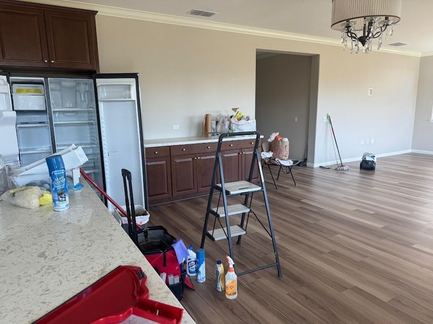 Empty kitchen/living room with new flooring. Refrigerator open, ladder, cleaning supplies, and cabinets visible.