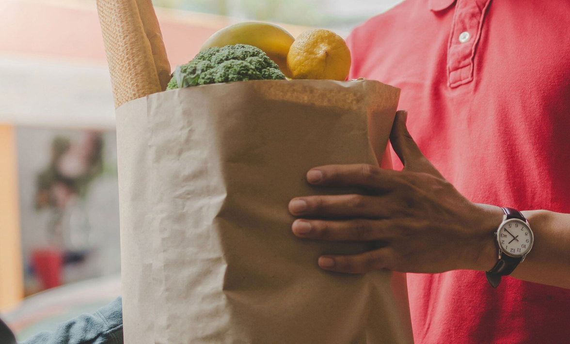 A man is holding a paper bag filled with vegetables and bread.