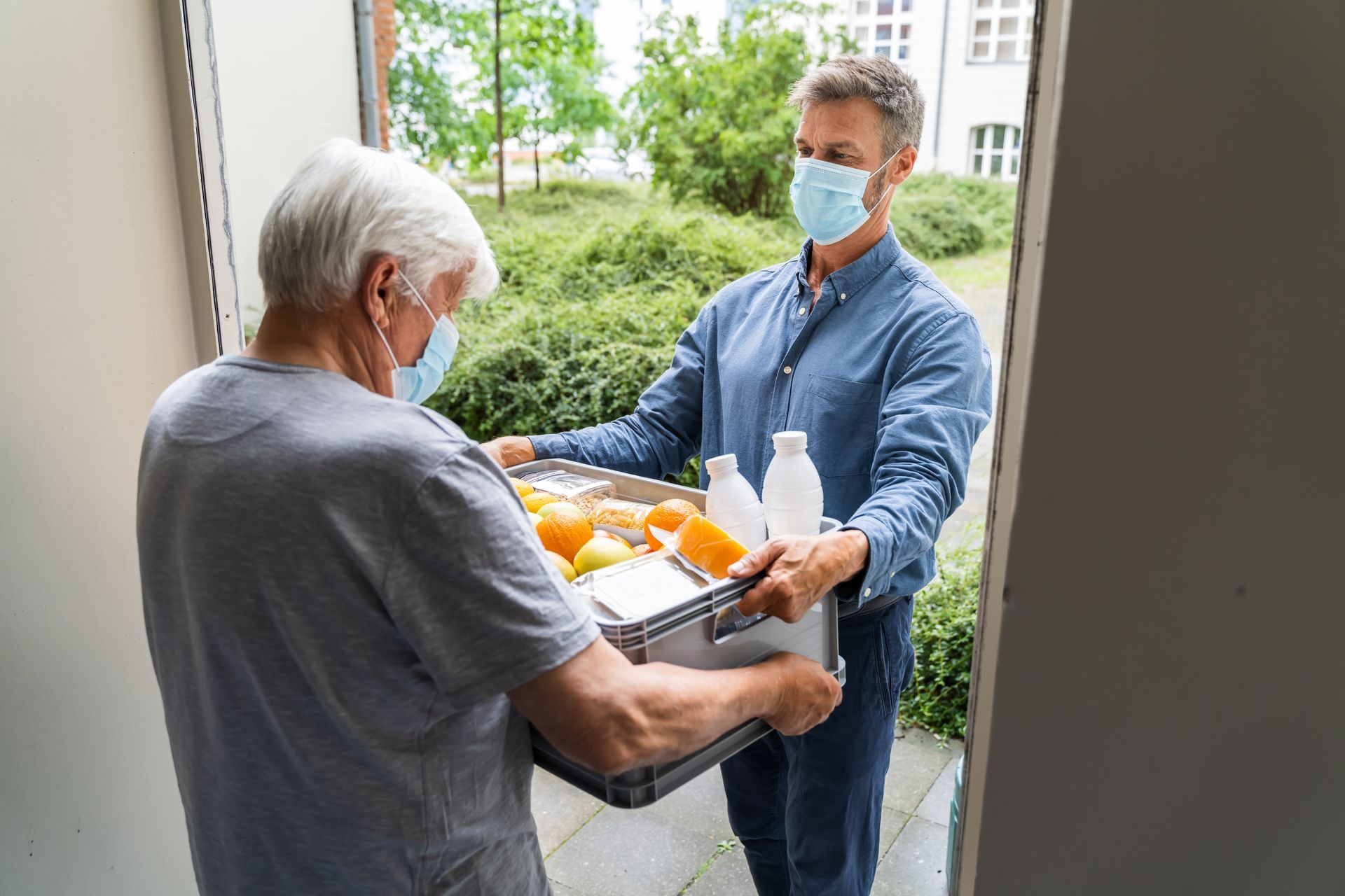 A man wearing a mask is delivering food to an elderly man.