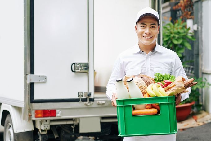 A delivery man is holding a green crate full of groceries in front of a truck.