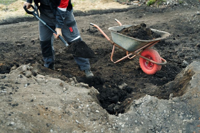 A person is digging in the dirt next to a wheelbarrow.
