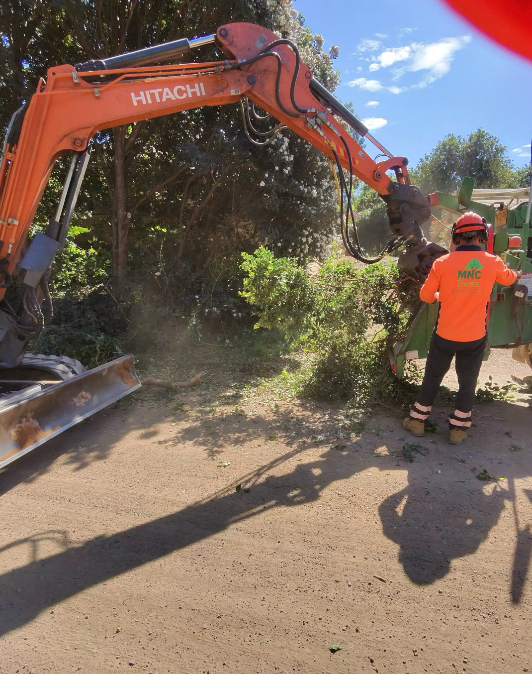 An orange Hitachi excavator feeding brush into a wood chipper, operator in safety gear. — MNC Trees in Port Macquarie, NSW