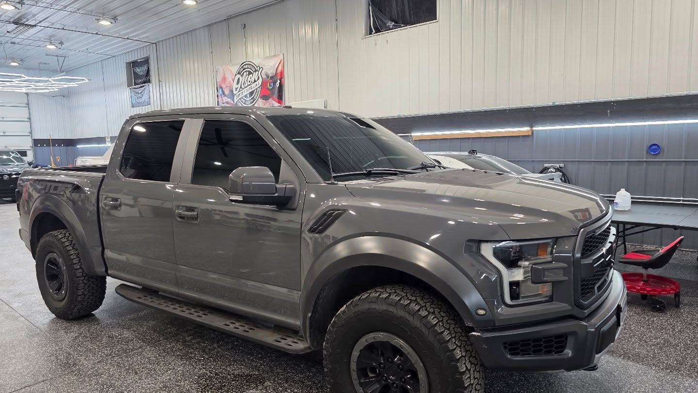A man applies protective film to the front of a gray Acura. He is kneeling near the car, with spray bottle.