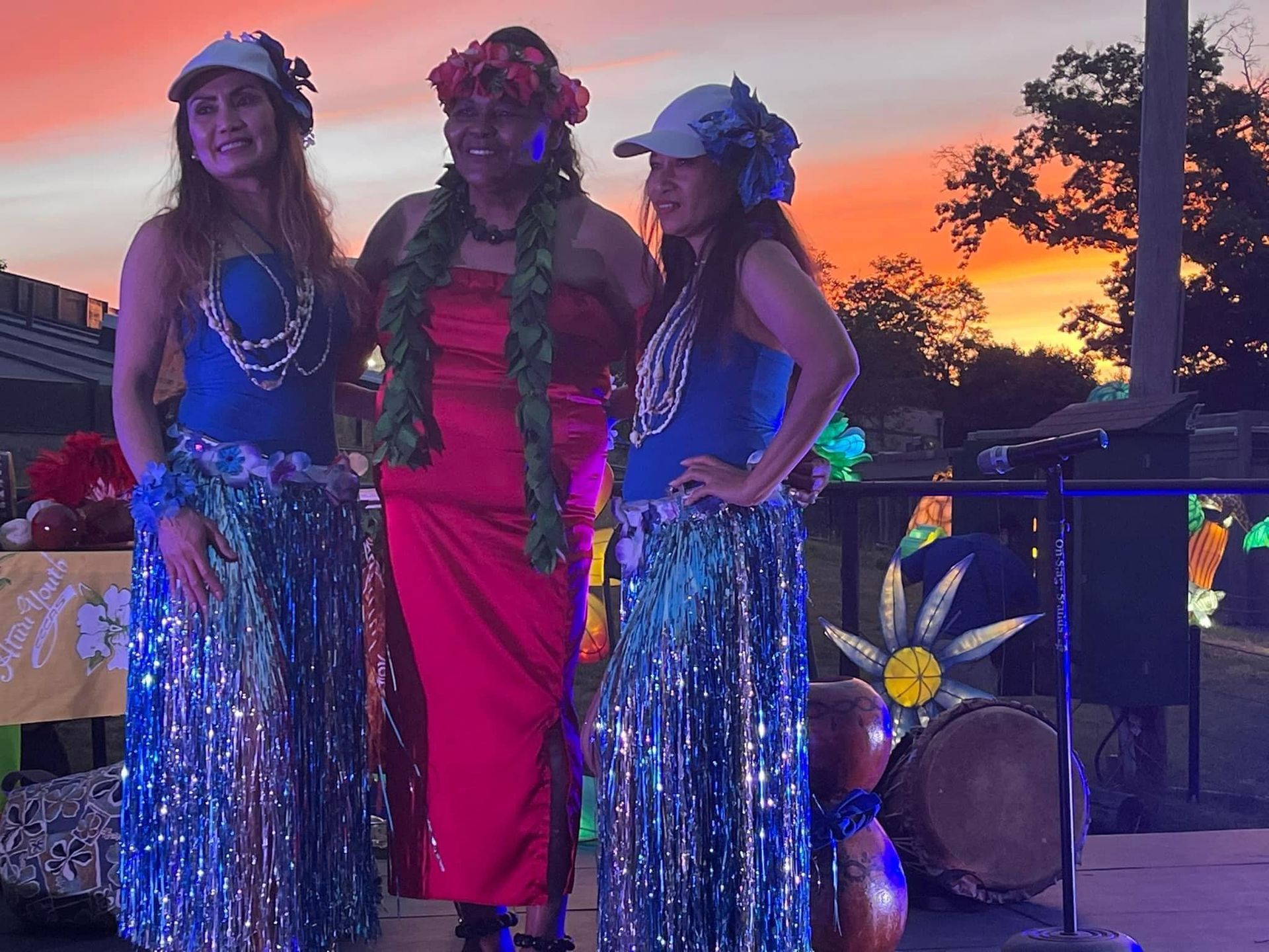 Three women in hawaiian costumes are standing next to each other in front of a sunset.
