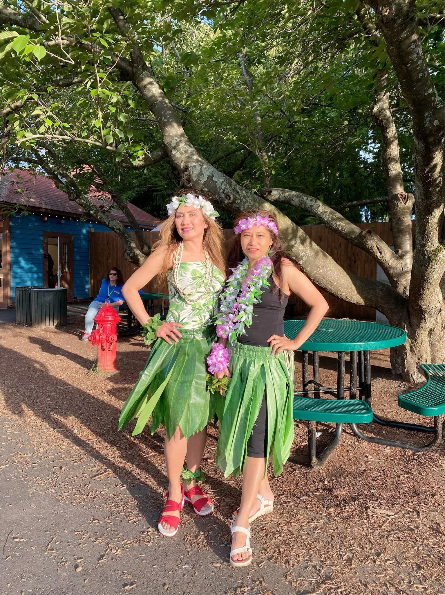 Two women in hawaiian costumes are standing next to each other in front of a picnic table.