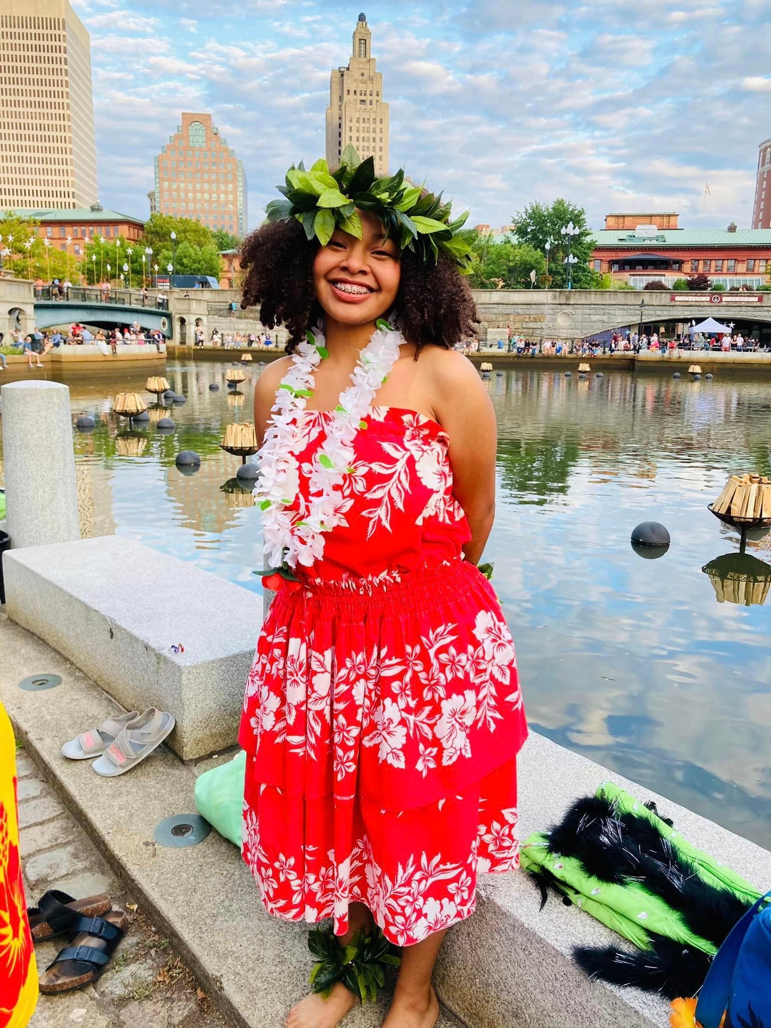 A woman in a red dress and a lei is standing next to a body of water.