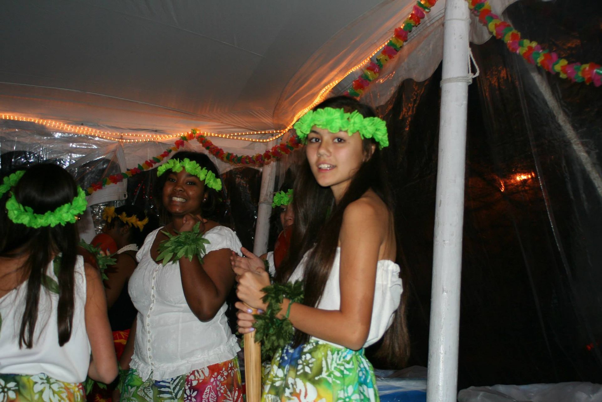 A group of women dressed in hawaiian costumes are standing under a tent.