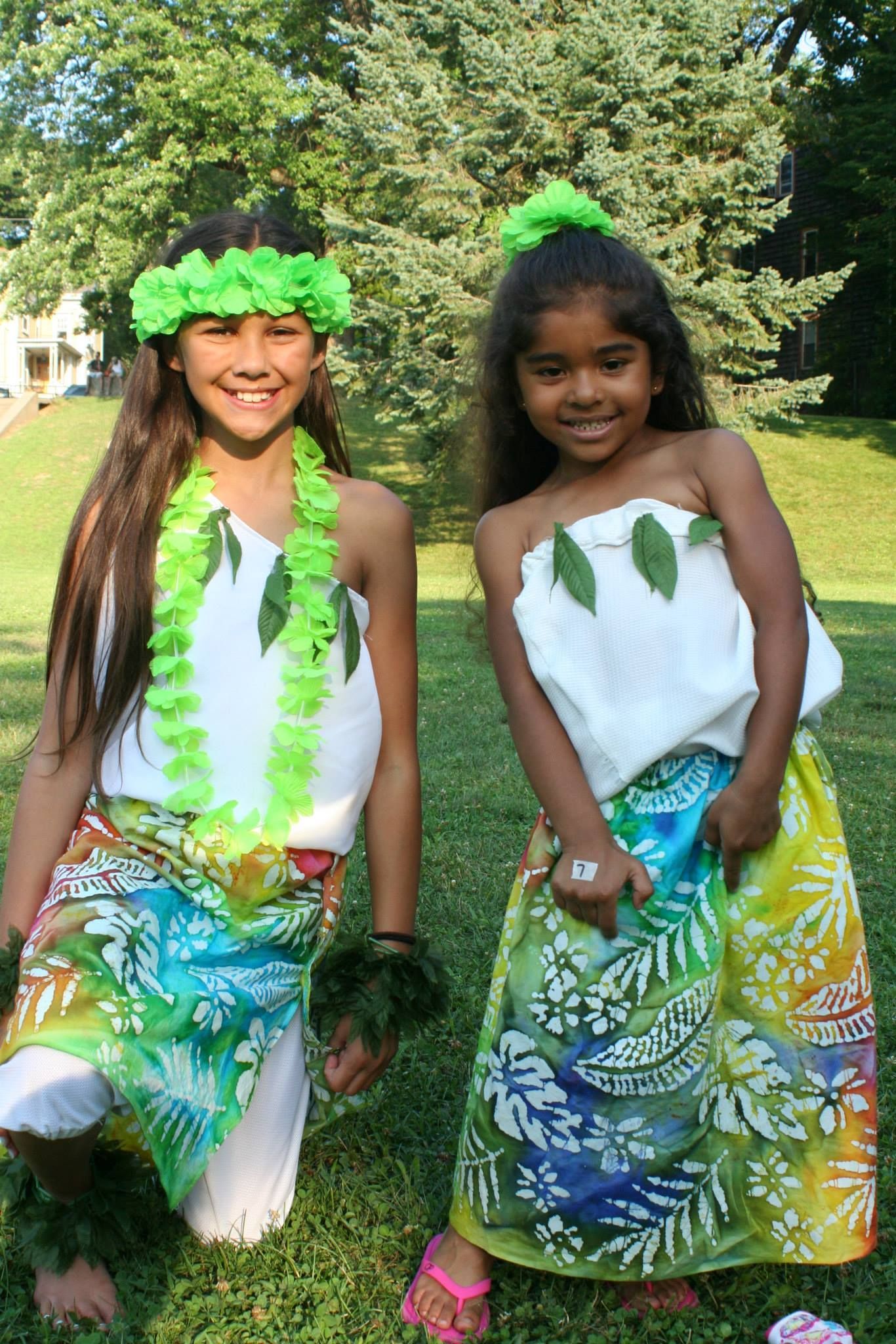 Two young girls dressed in hawaiian costumes are standing next to each other in the grass.