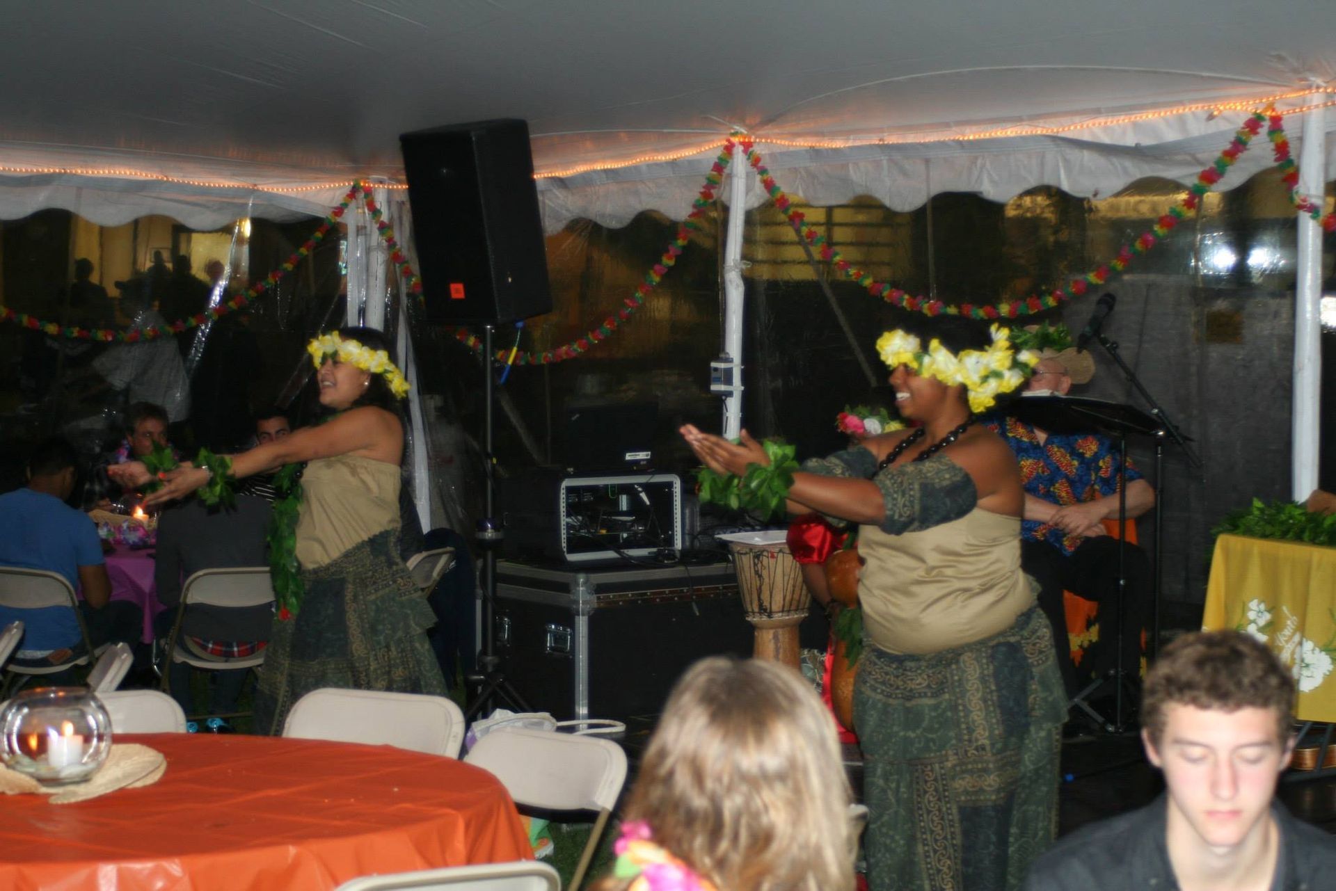 A group of people are sitting at tables under a tent watching hula dancers perform