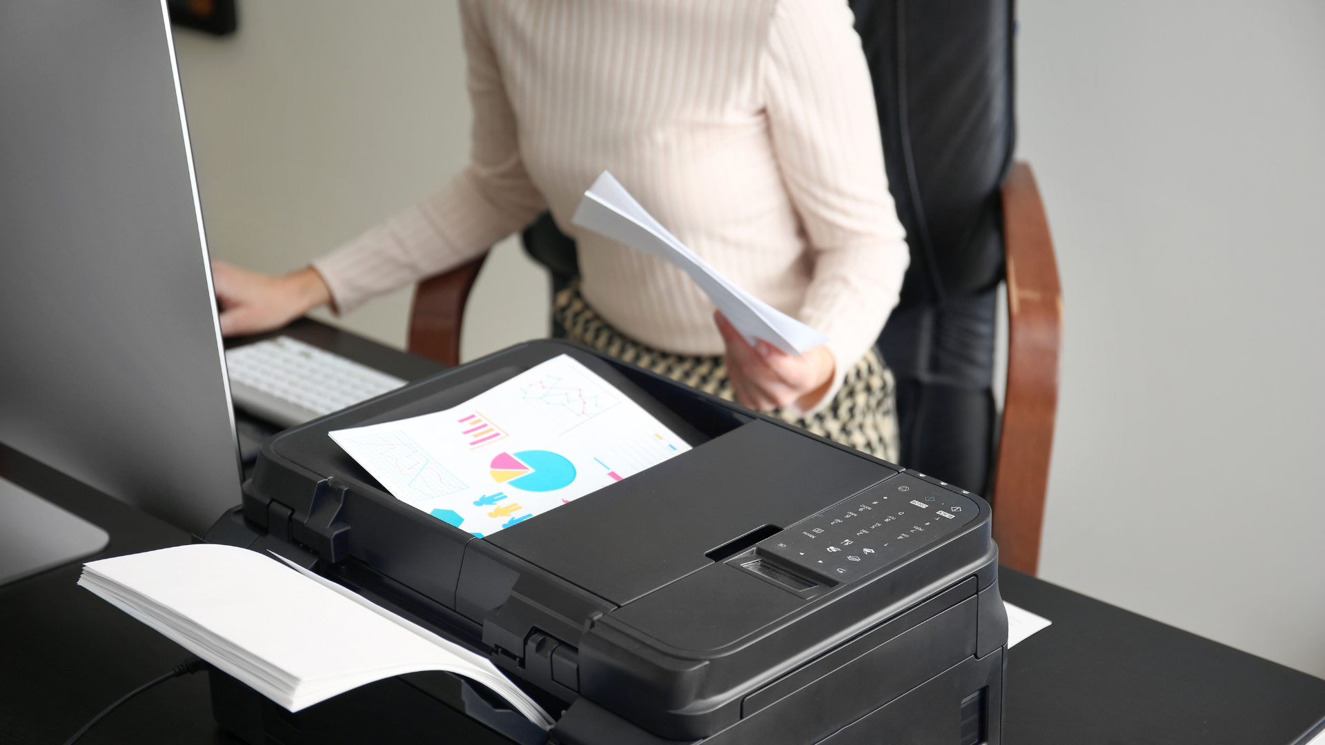 Person printing documents at an office desk; printer in foreground, computer in background.