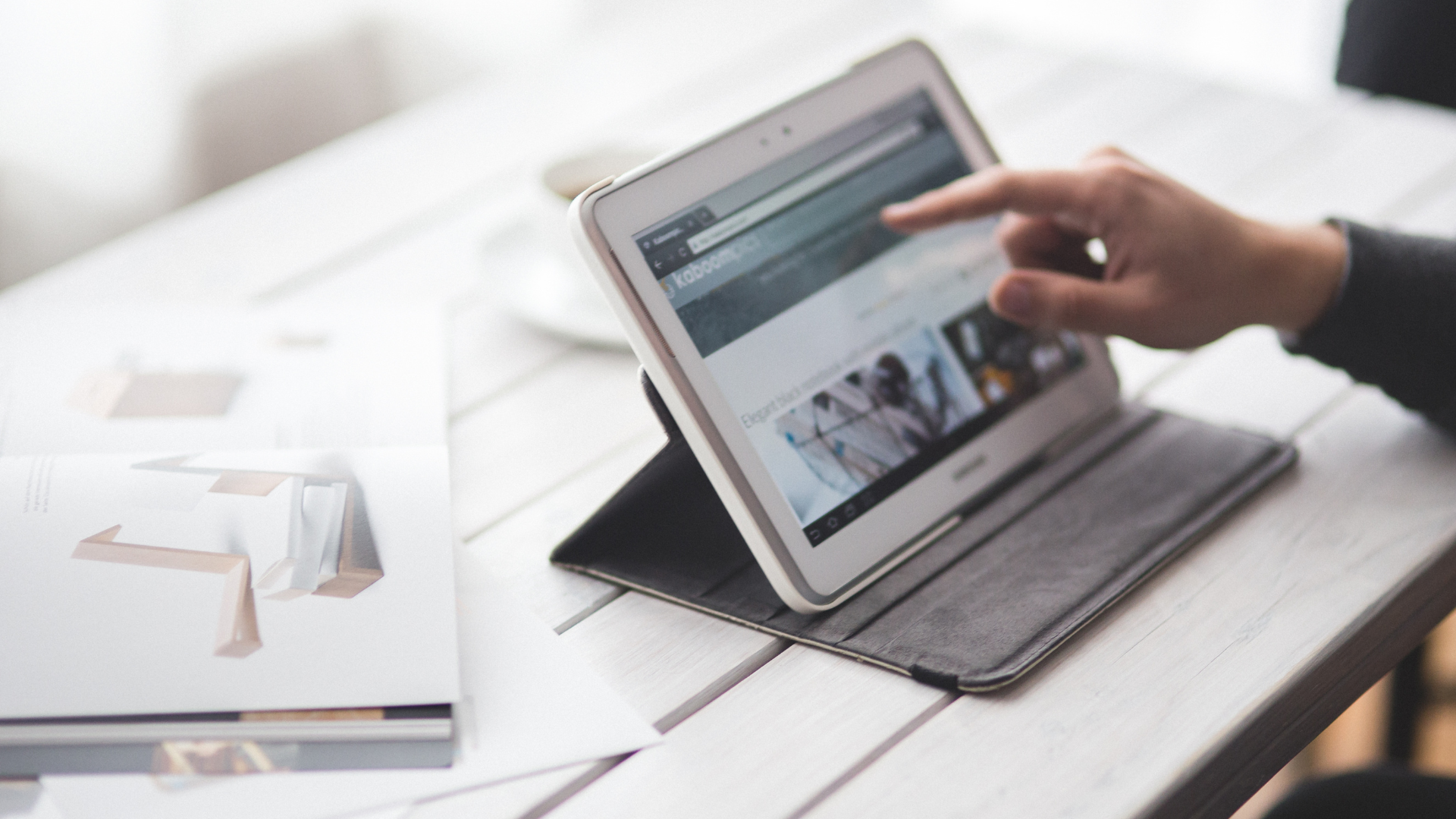 Person's hand pointing at a tablet screen on a white wooden table. Open webpage, coffee cup and book nearby.