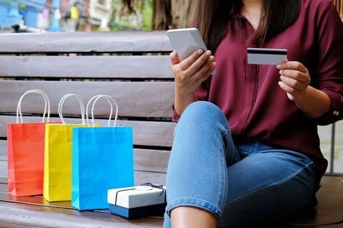 Woman on bench, shopping online with phone and credit card, colorful bags nearby.