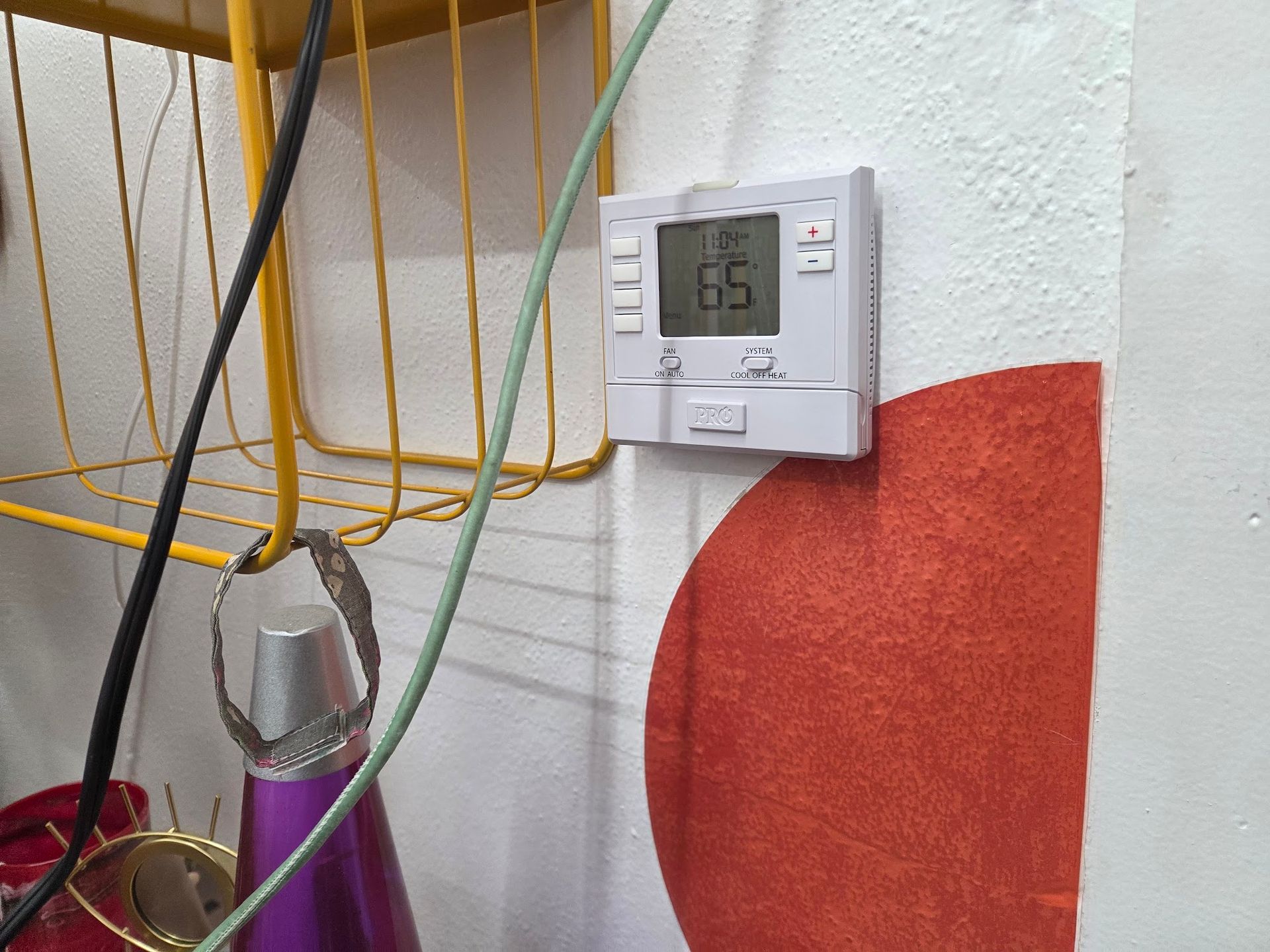 Thermostat on white wall, set to 65 degrees. Red heart shape and yellow shelf with cords are visible.
