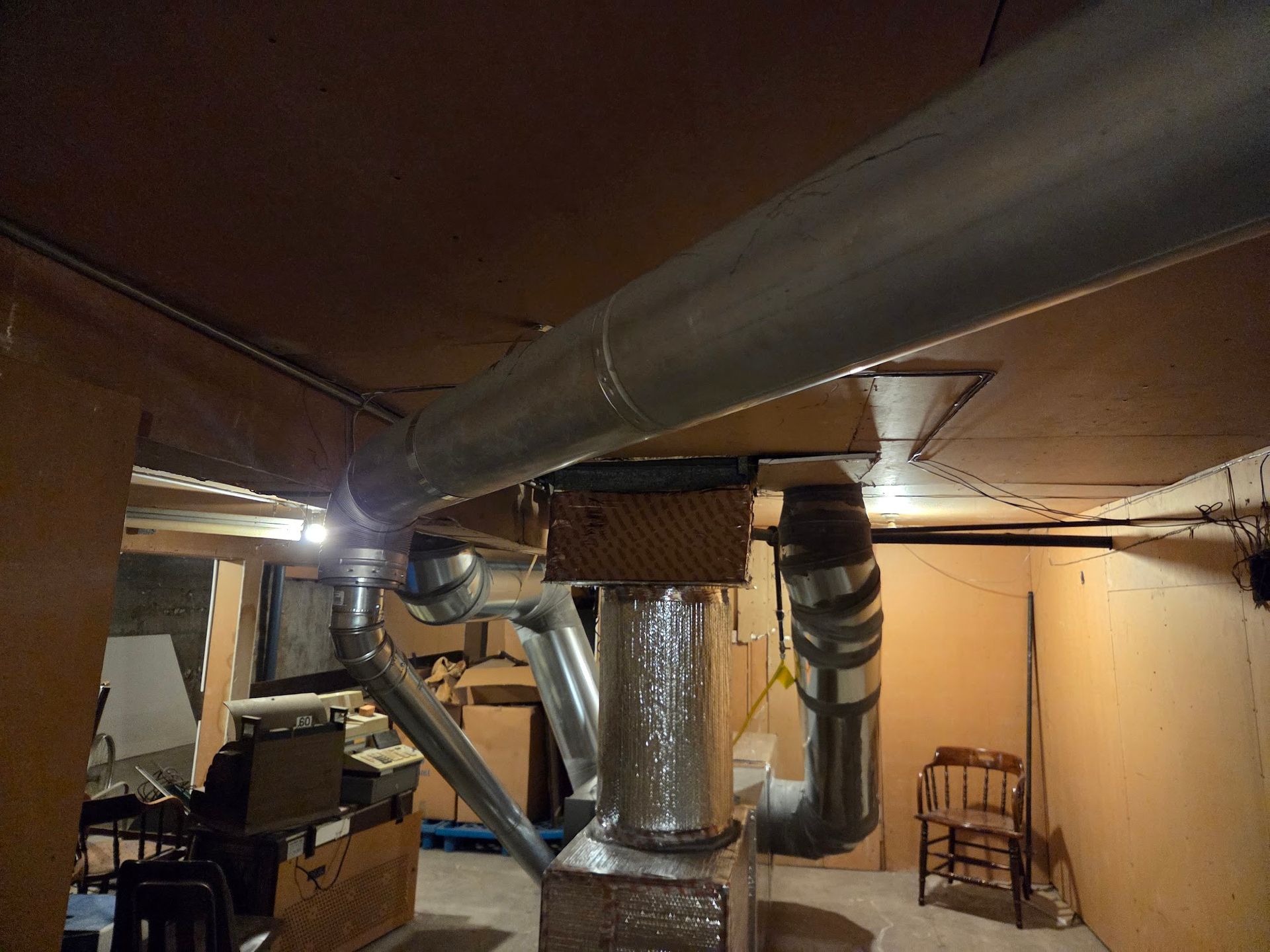 HVAC ductwork in a basement ceiling, with insulated and uninsulated metal pipes. Brown walls and a chair are visible.