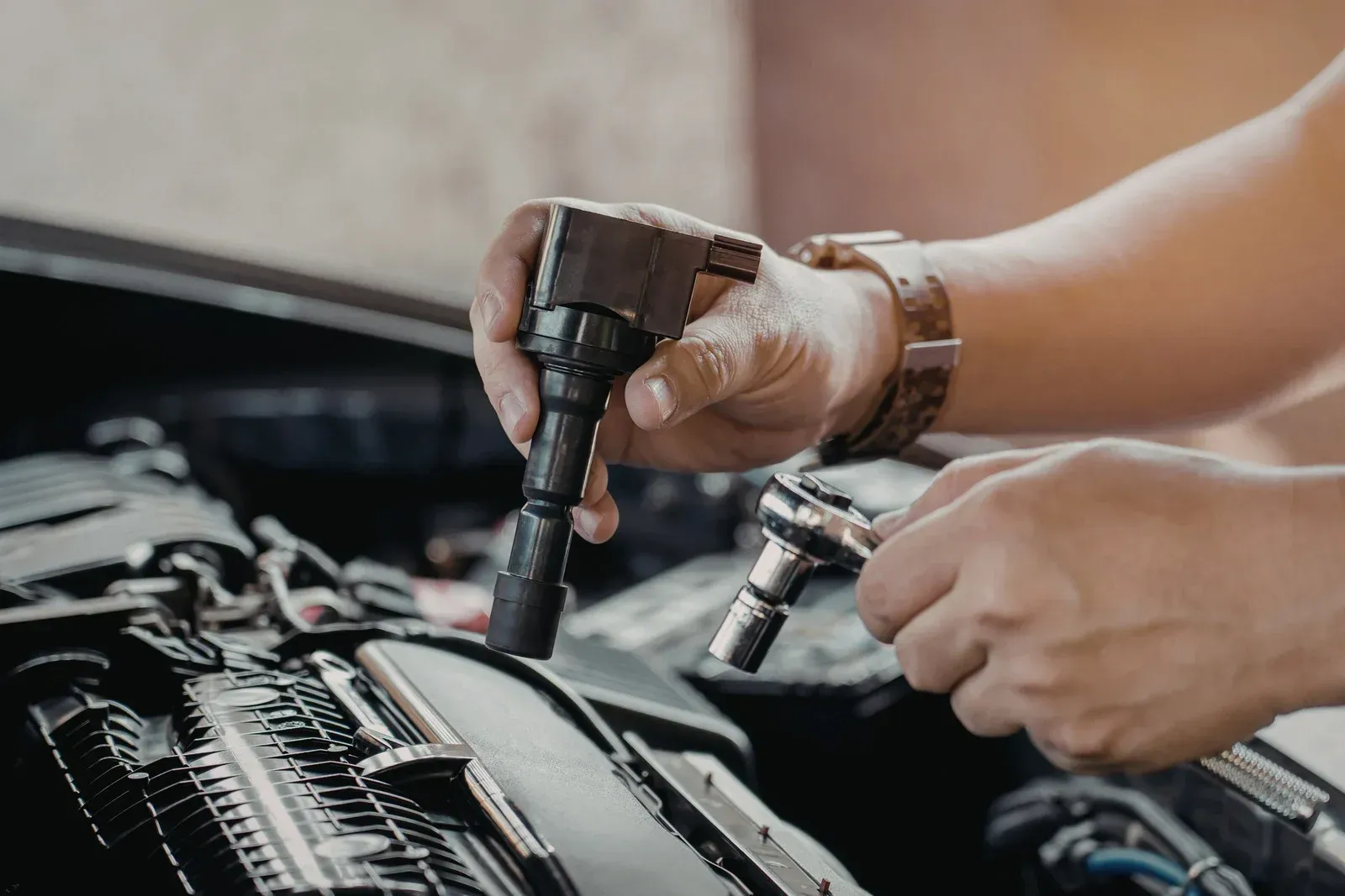 Mechanic's hands holding an ignition coil and a wrench while working on a car engine.