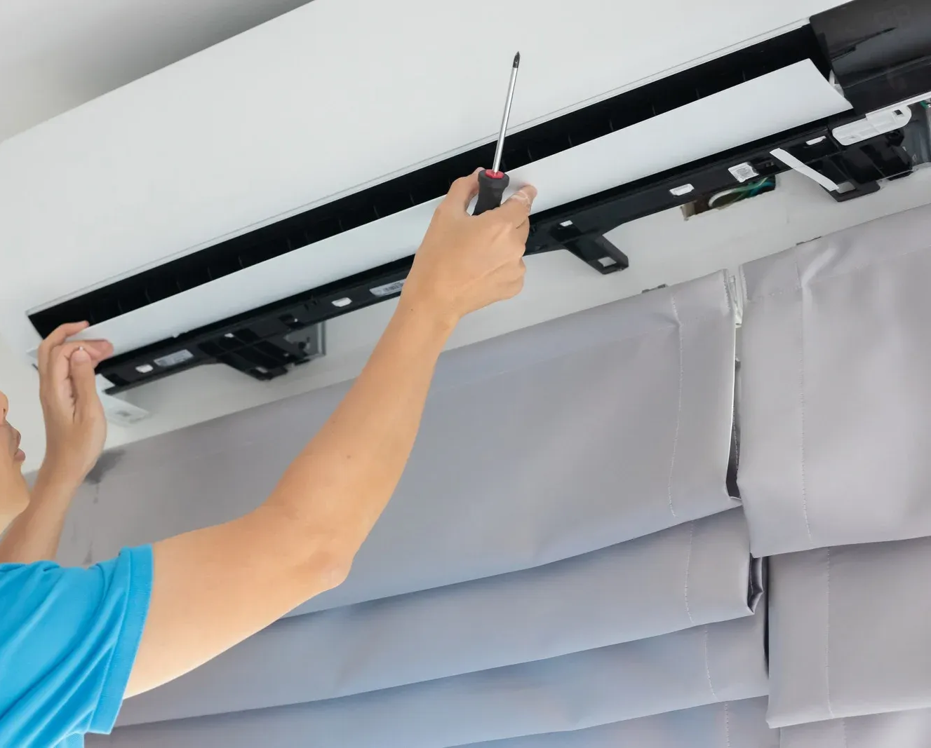 Person repairs an air conditioner using a screwdriver. White ceiling, gray blinds, close-up.
