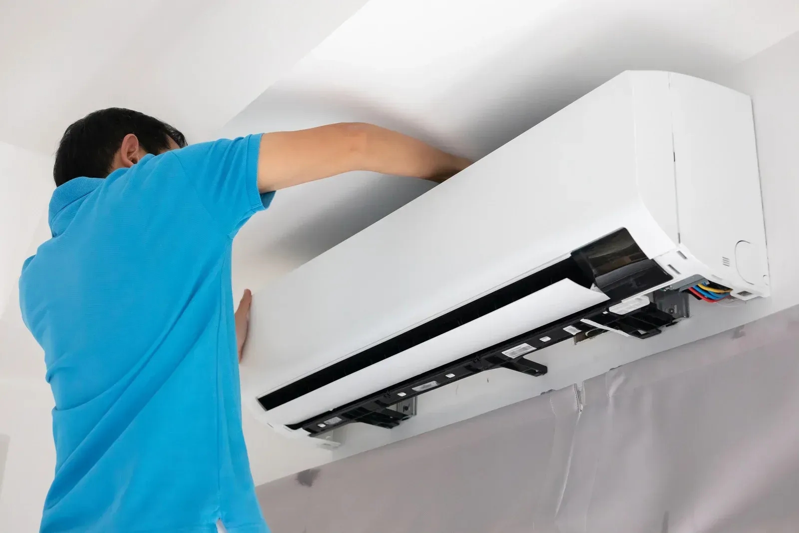 Man in blue shirt installing a white air conditioning unit on a white wall.