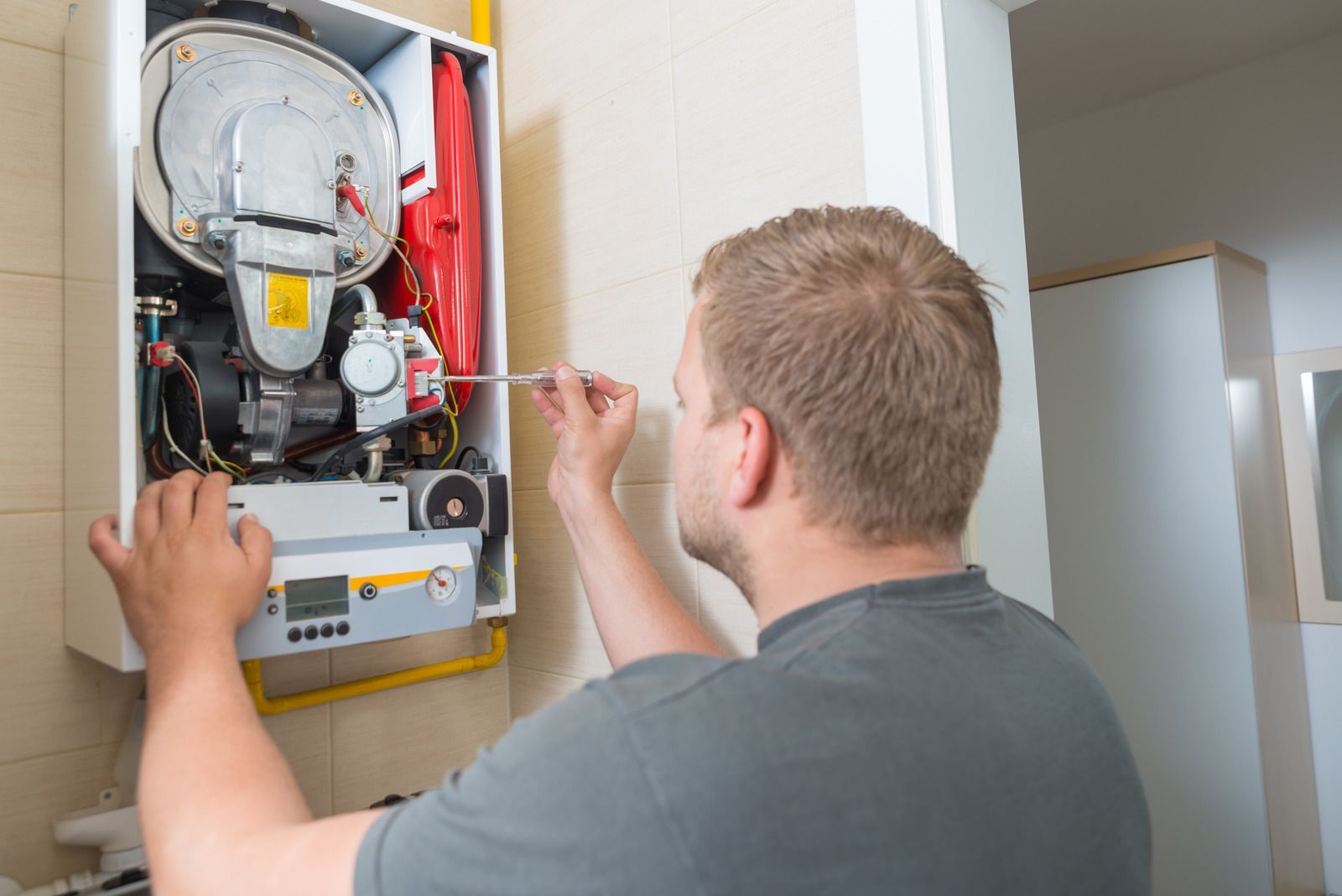 Person repairs a white boiler on a wall, using a screwdriver.
