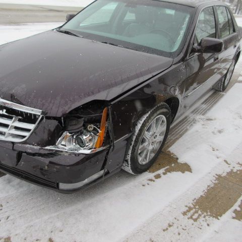 Damaged burgundy Cadillac sedan on snow-covered ground; front-end collision with broken headlight.