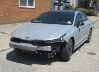 Gray Kia sedan with front-end damage; parked on a city street. Black roof, missing bumper.