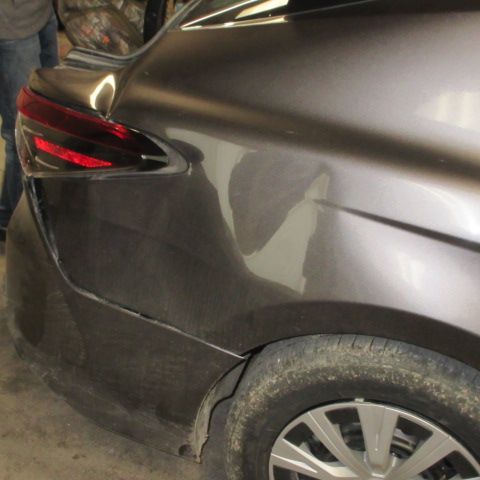 A gray car with a dent in the rear panel, near the taillight and tire.