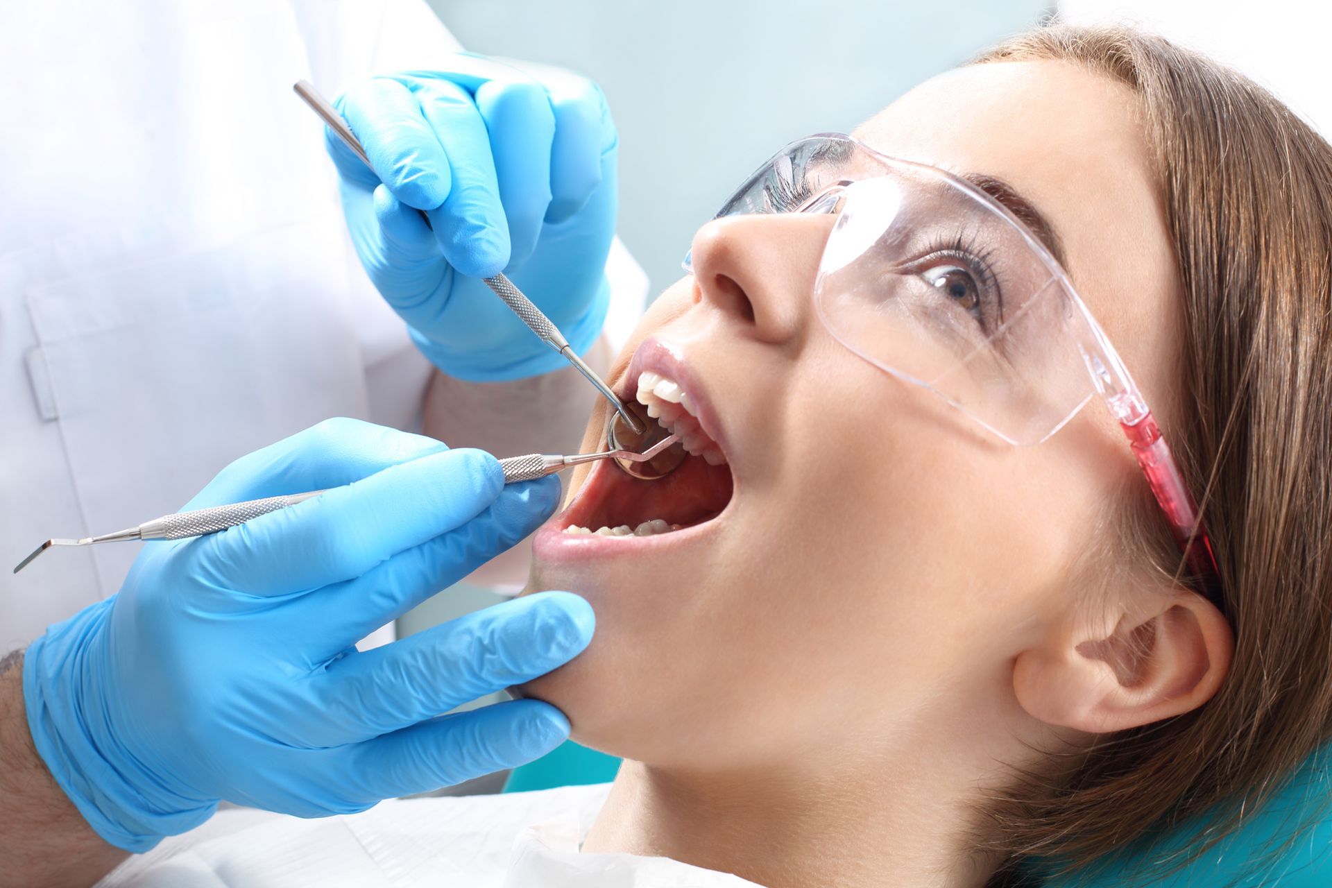Dentist examining a woman's teeth with tools, wearing blue gloves and eye protection.
