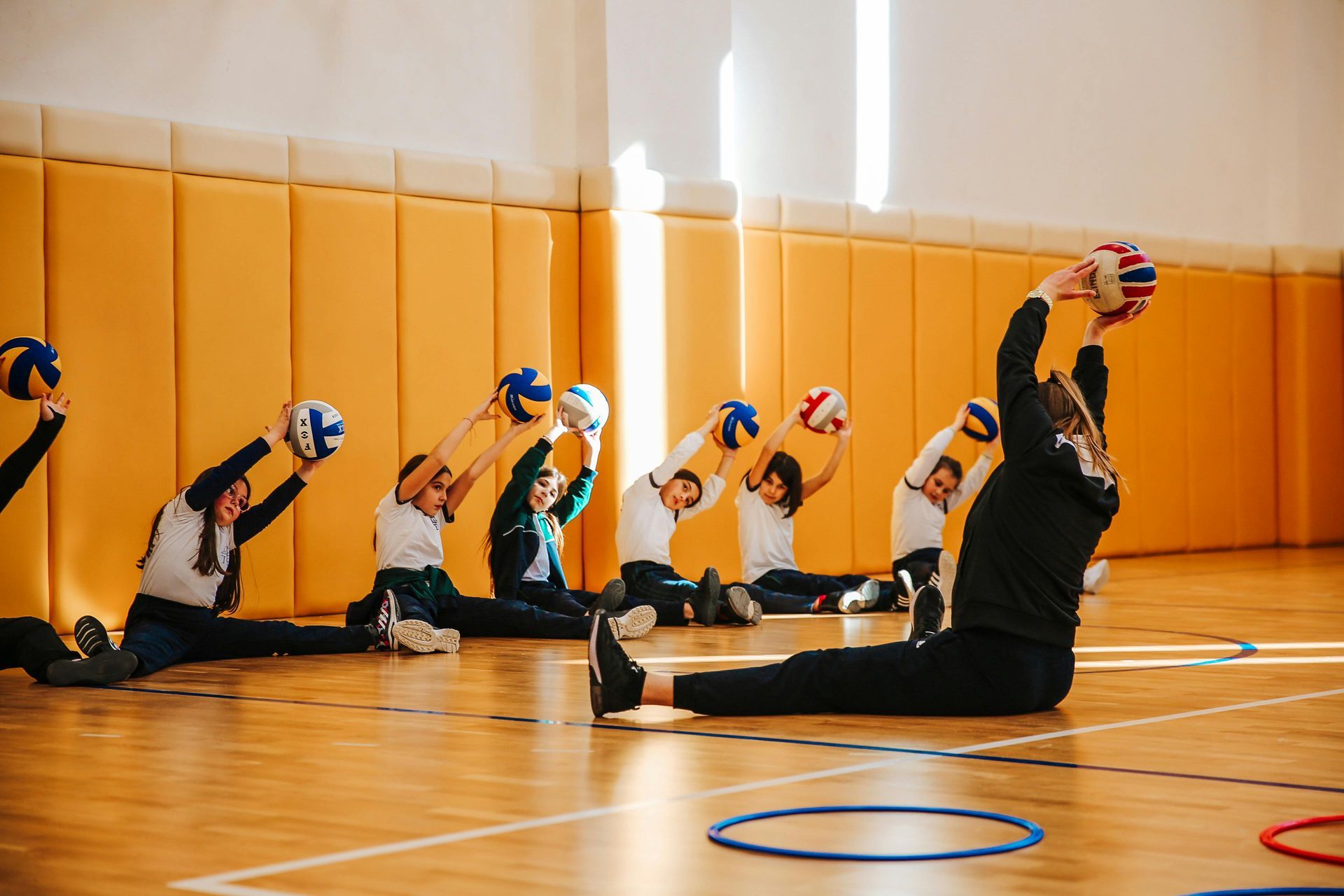Children in gym class stretching with balls, raising arms.