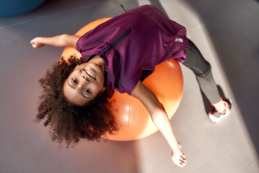 Young girl with curly hair smiles while lying on an orange exercise ball, reaching arms out.