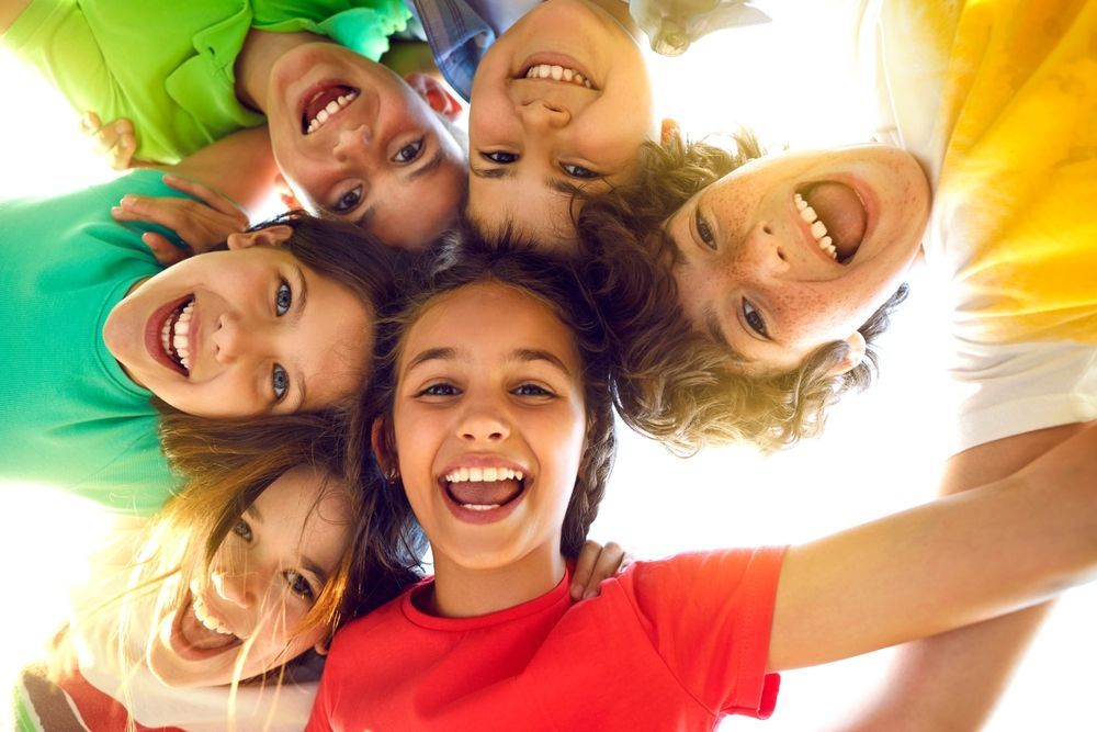 Group of smiling children in a huddle, looking down. Bright colors and happy expressions.