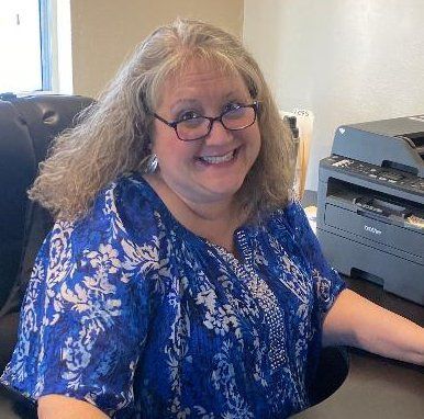 A woman with white curly hair with specs and blue dress is smiling while sitting in office