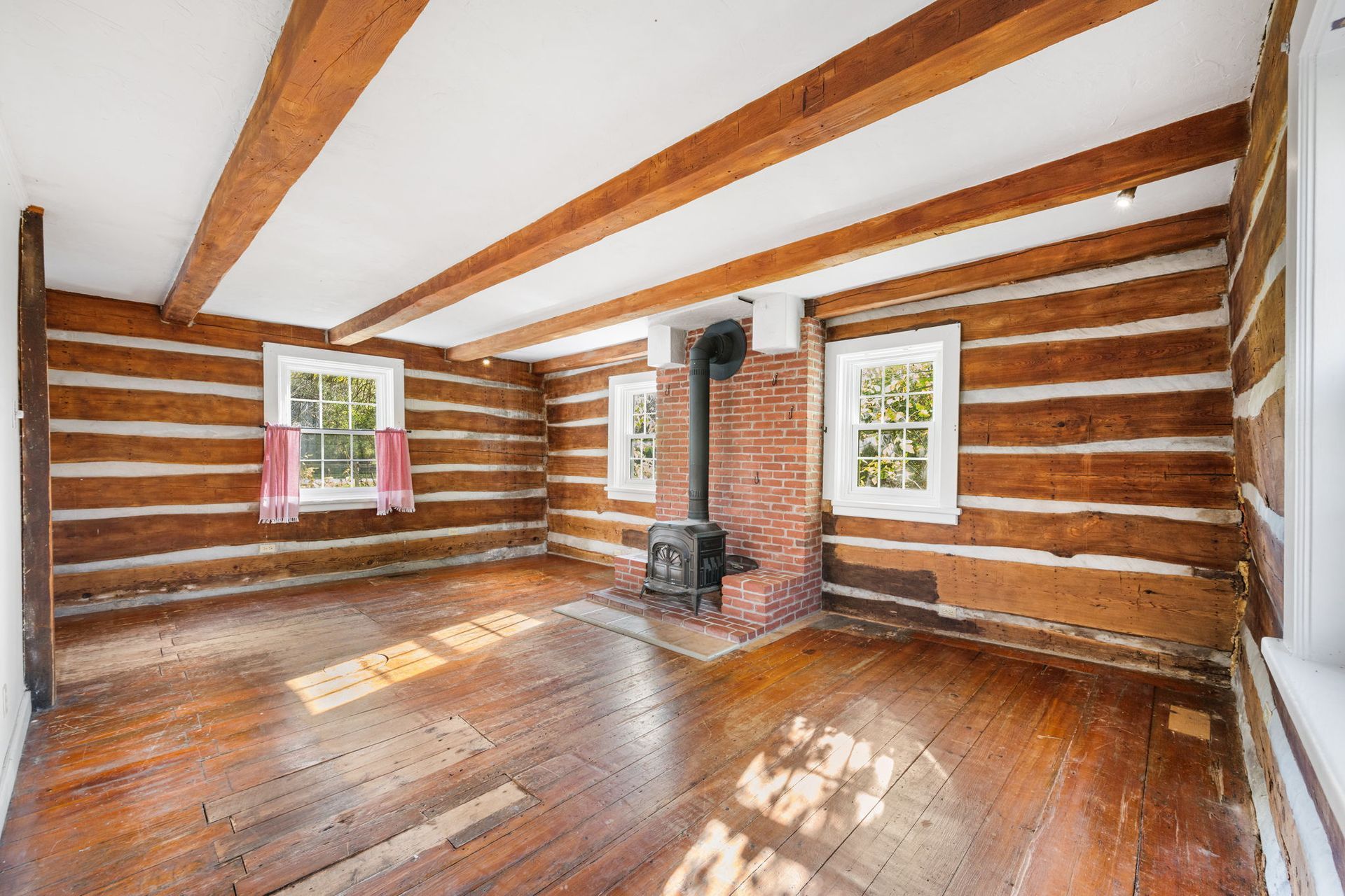 Interior of a cabin with wooden walls, floor, and ceiling beams. A brick chimney and wood stove are in the center.