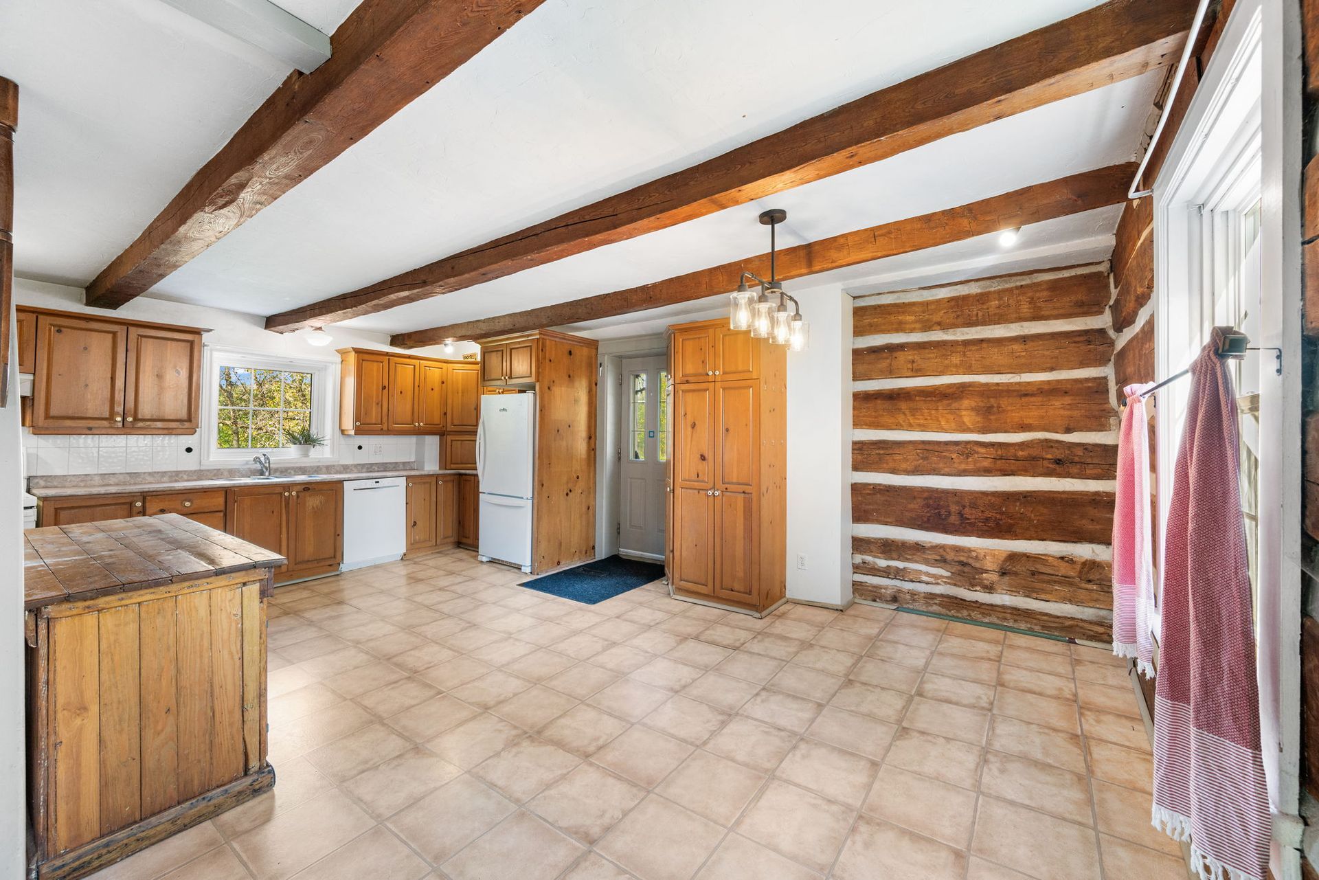 Kitchen with wooden beams, cabinets, and a log wall. Beige tile floor and window with towels.